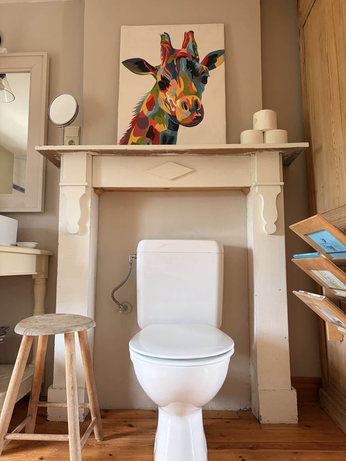 A bathroom with a white toilet, a wooden stool, a mirror, and a colorful giraffe painting above the toilet.