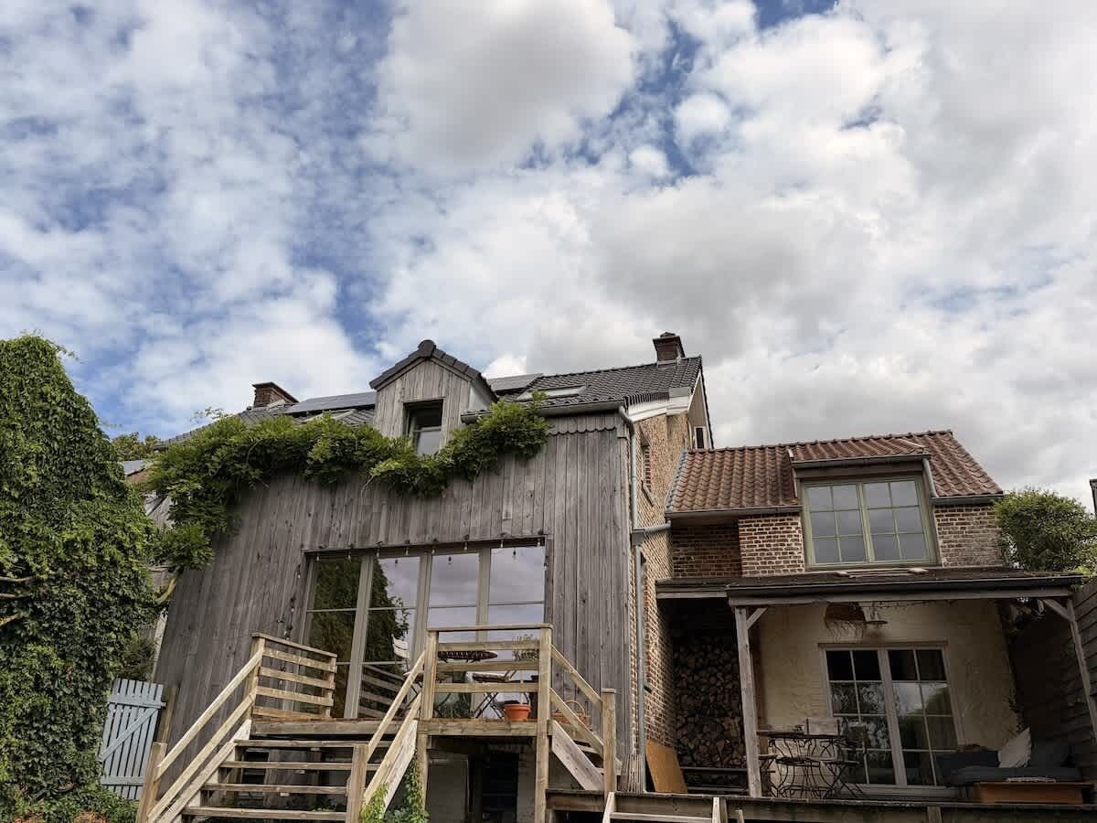 Una casa de dos pisos con una escalera de madera que conduce a una puerta, rodeada de follaje.