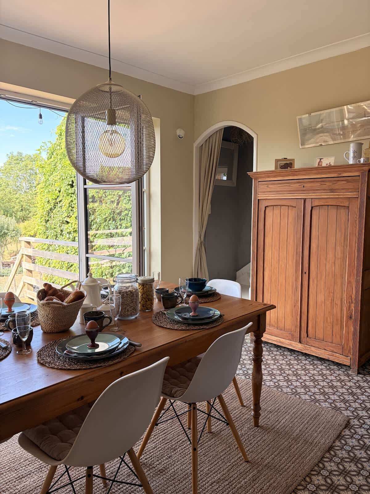 Uma sala de jantar com uma mesa de madeira posta para quatro pessoas, cadeiras, um lustre e vista para o verde lá fora.
