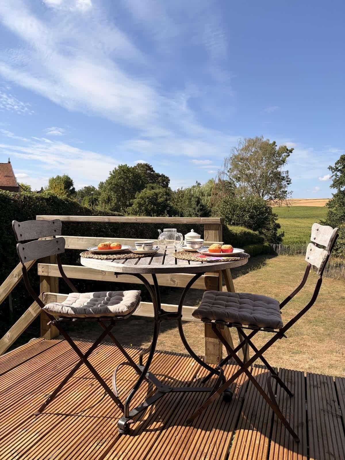 Un patio esterno in legno con un tavolo rotondo e sedie, un boccale, tazze e un piatto con cibo.