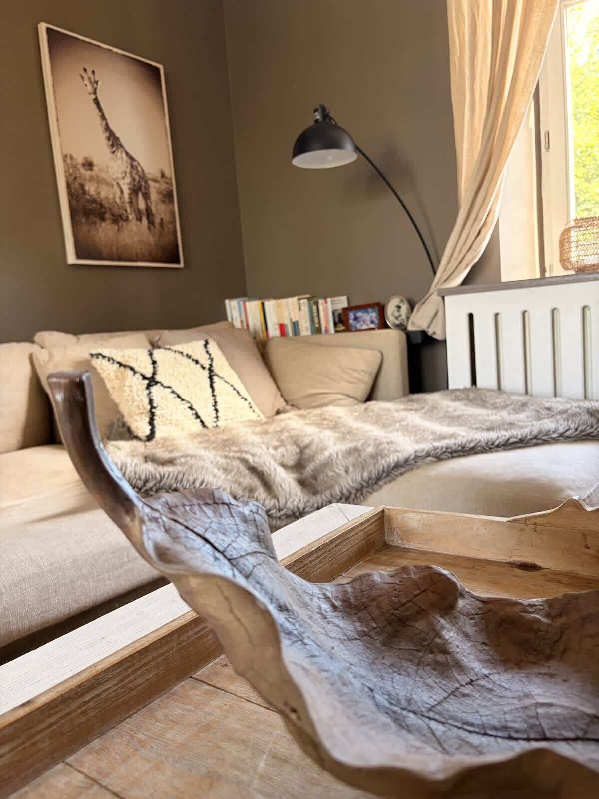Ein Zimmer mit einer Couch, einem Holztischchen, einer Lampe, einem Heizkörper und einem Fenster mit Vorhängen.