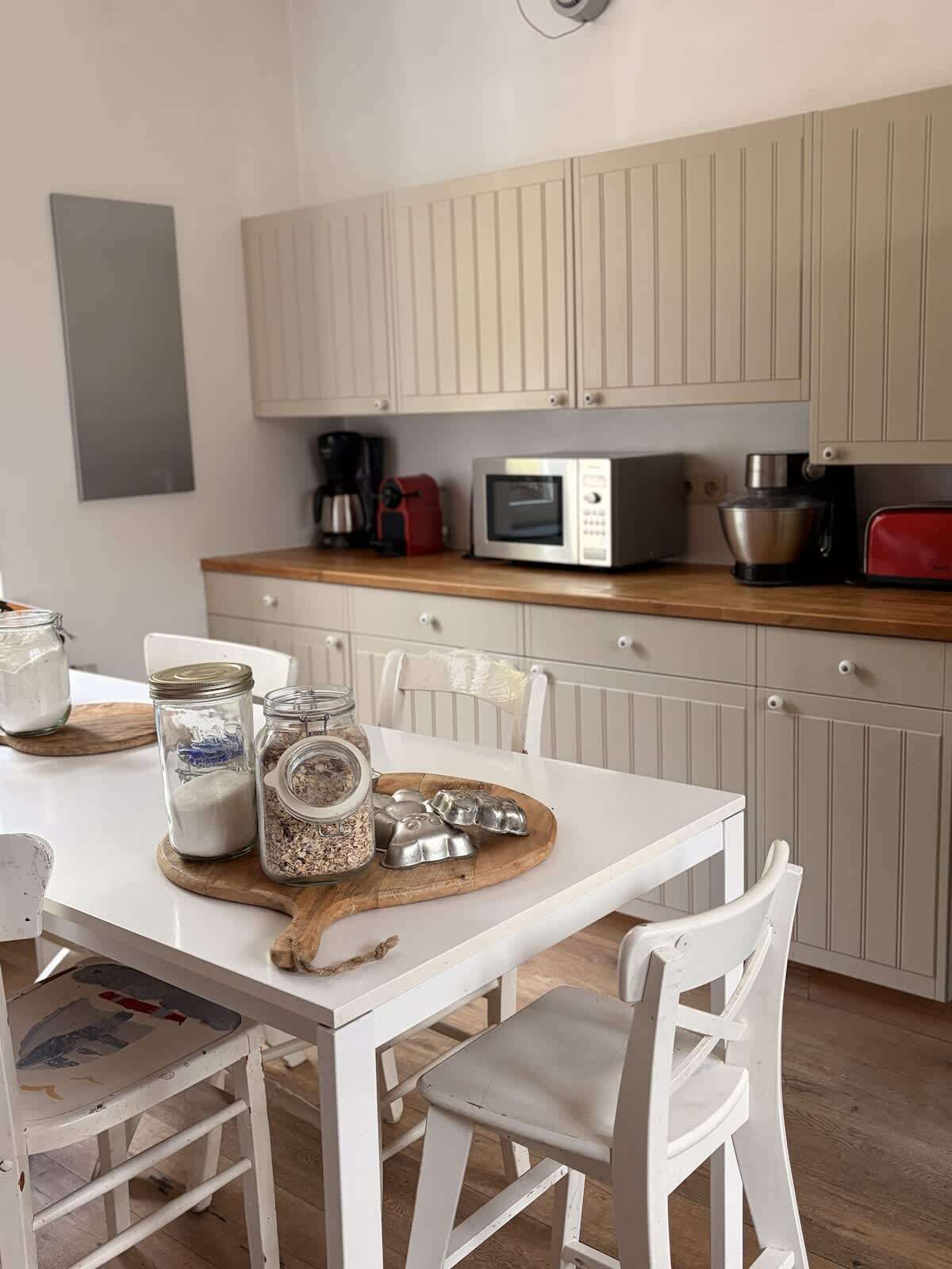 Una cocina con armarios blancos, microondas, cafetera y un tostador rojo. Hay dos sillas blancas y una mesa de madera con frascos y moldes metálicos para muffins.