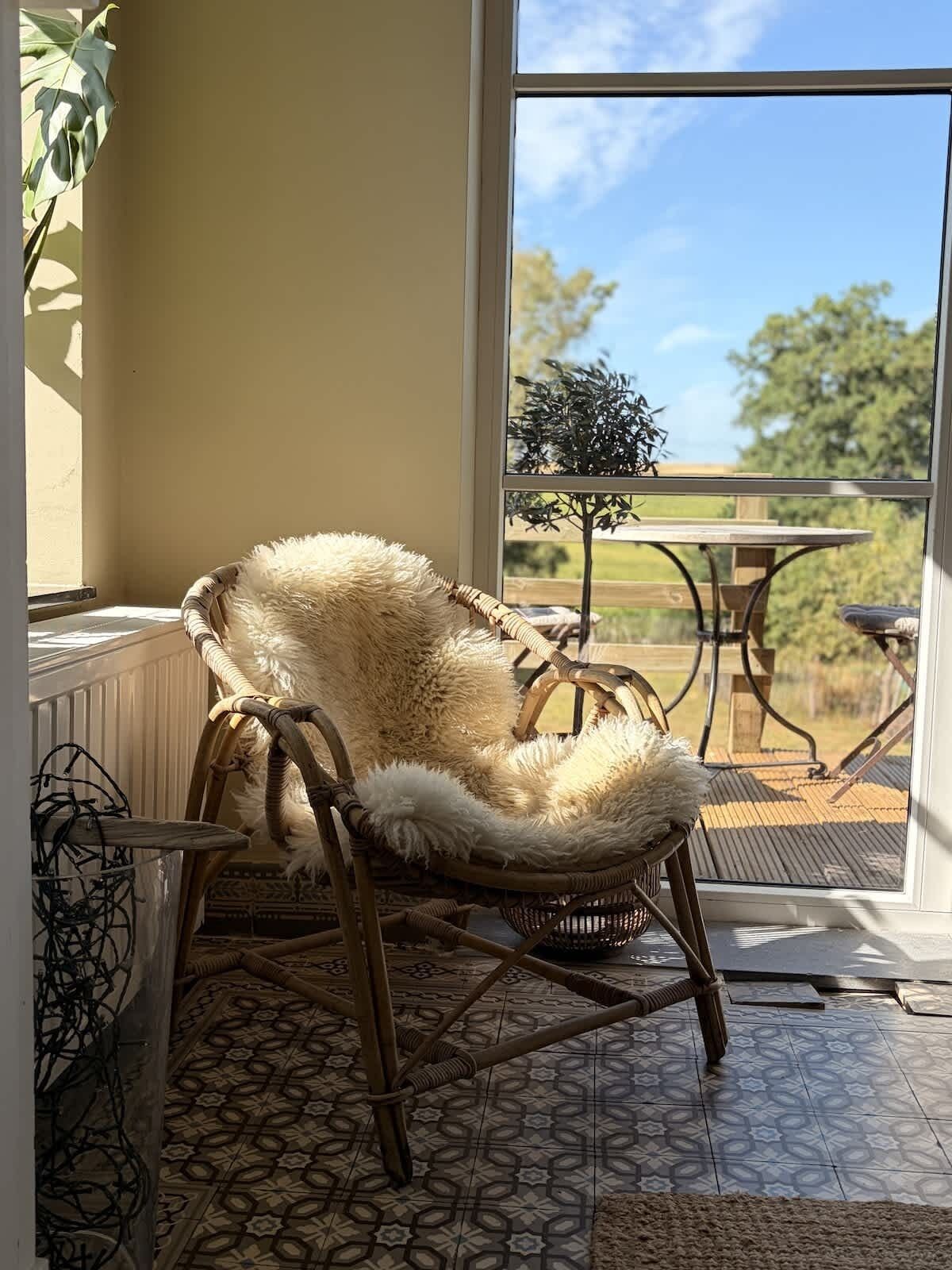 Una silla de mimbre con un cojín mullido en una habitación con baldosas estampadas, vista a un patio con una mesa y sillas, y una planta en maceta.