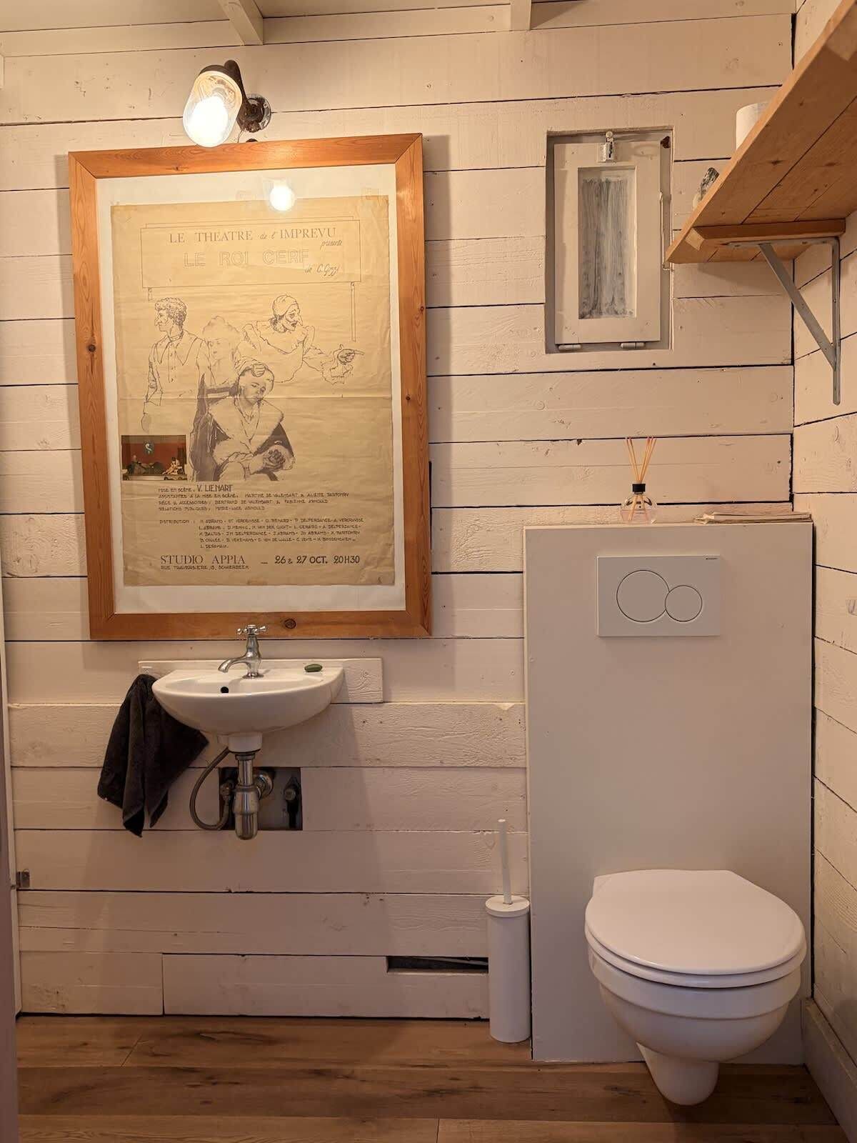 A bathroom with a wooden-framed poster, a sink, a toilet, and a small shelf with a bottle.