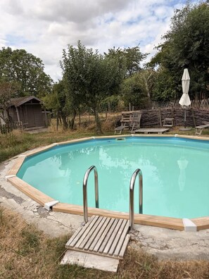 A swimming pool with a wooden deck and metal ladder, surrounded by trees and a wooden fence. - Charmant cottage à la campagne (Jodoigne)