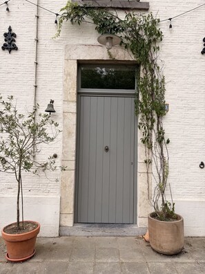 A white brick building with a grey door, a small potted plant, and a string of lights.