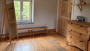 Una stanza con pavimento in legno, una panca, un comò di legno, una porta e una finestra con vista sul verde.