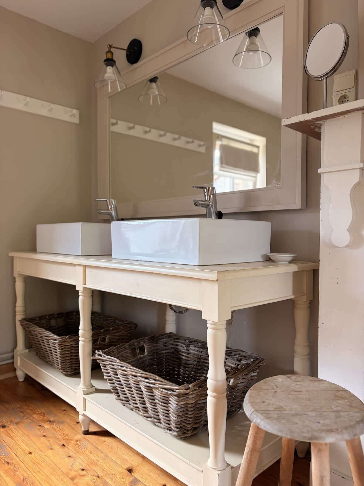 Une salle de bain avec un meuble-lavabo blanc, un grand miroir et des paniers en osier sur des étagères.