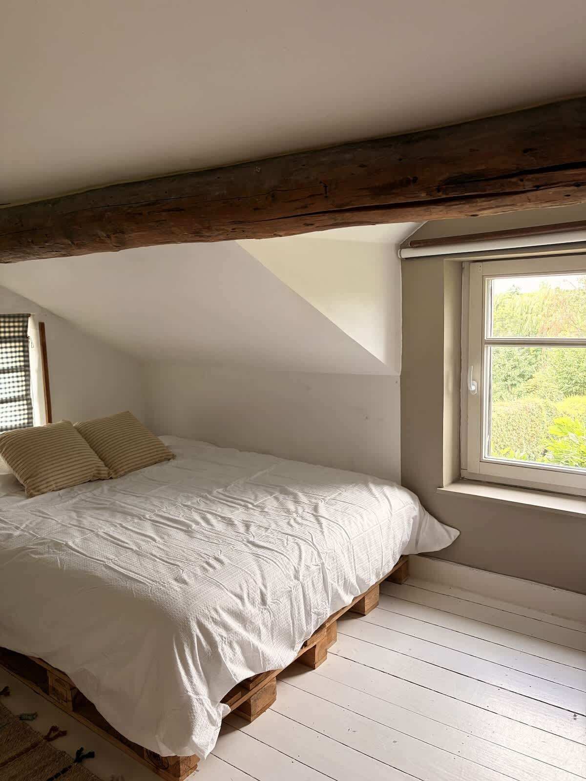 Une chambre avec un plafond en pente, une fenêtre donnant sur un paysage verdoyant et un lit recouvert d’une couverture blanche et agrémenté de deux oreillers.