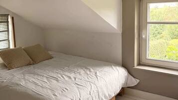 Une chambre avec un plafond en pente, une fenêtre donnant sur un paysage verdoyant et un lit recouvert d’une couverture blanche et agrémenté de deux oreillers.