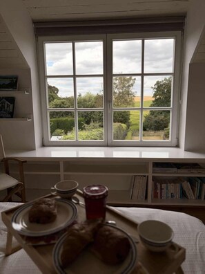 A room with a large window overlooking a garden, a bookshelf, and a table with plates and cups. - Charmant cottage à la campagne (Jodoigne)