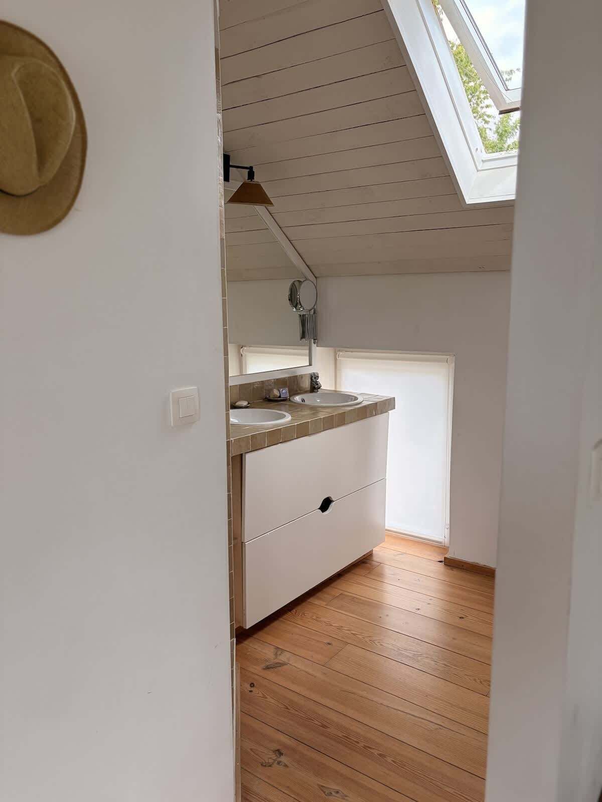 Een smalle gang die leidt naar een badkamer met dakraam, houten vloer en een witte wastafel met spiegel.