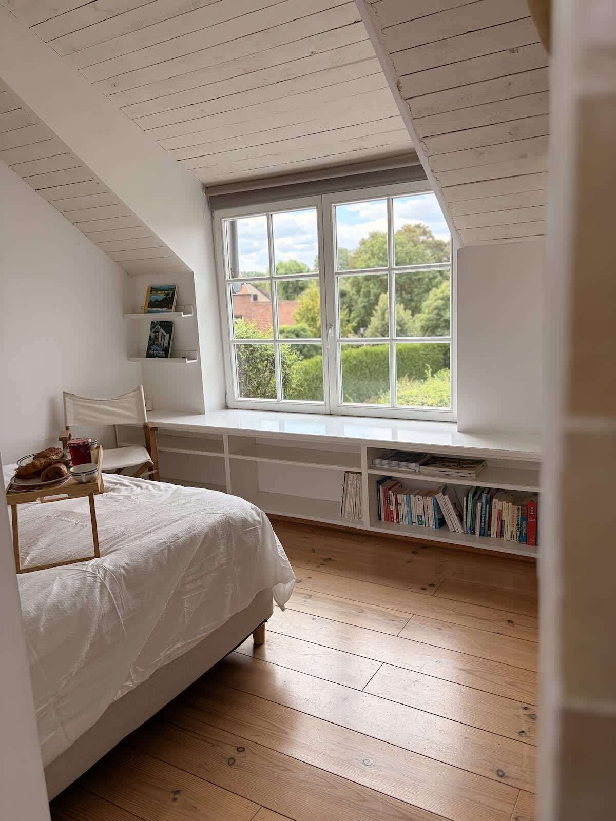 Een slaapkamer met een groot raam, een bed, een boekenkast en een klein tafeltje met boeken en een beker.