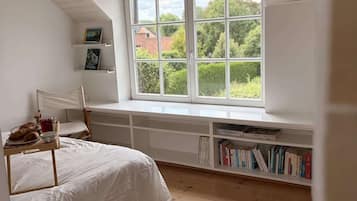 Een slaapkamer met een groot raam, een bed, een boekenkast en een klein tafeltje met boeken en een beker.