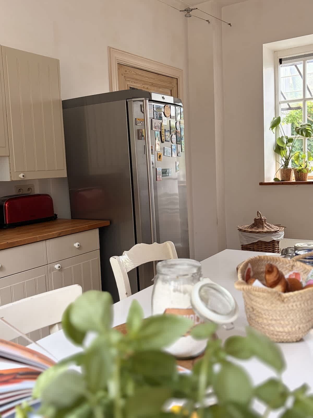 Une cuisine équipée d’un réfrigérateur, de meubles de rangement en bois, d’une table à manger avec des chaises et d’une fenêtre agrémentée de plantes.