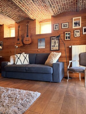 A cozy living room with a dark blue sofa, a wooden floor, and a ceiling with exposed beams.