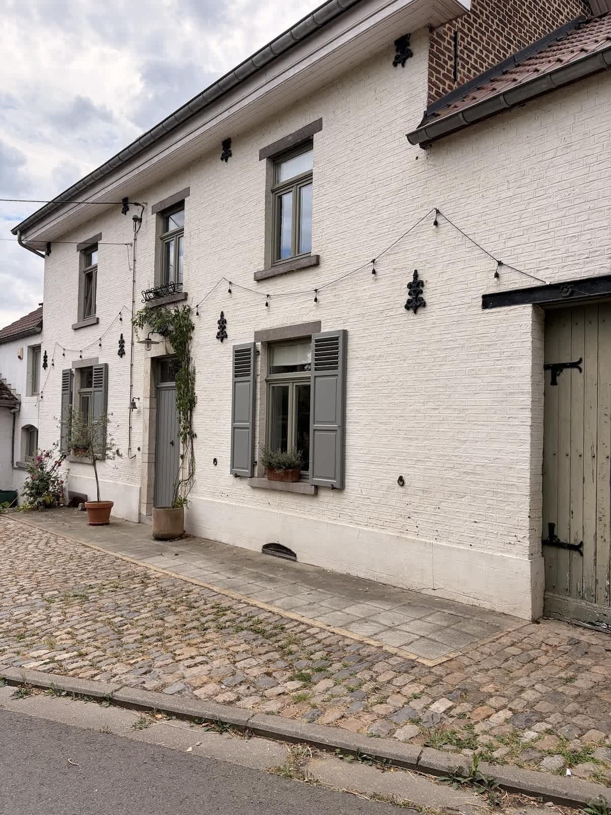 Ein weißes Backsteinhaus mit mehreren Fenstern, einer Tür und einem kleinen Gartenbereich mit Blumentöpfen.