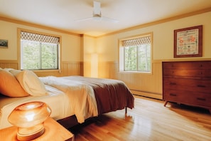 Quarto com cama, penteadeira, janelas e relógio fixado na parede.
