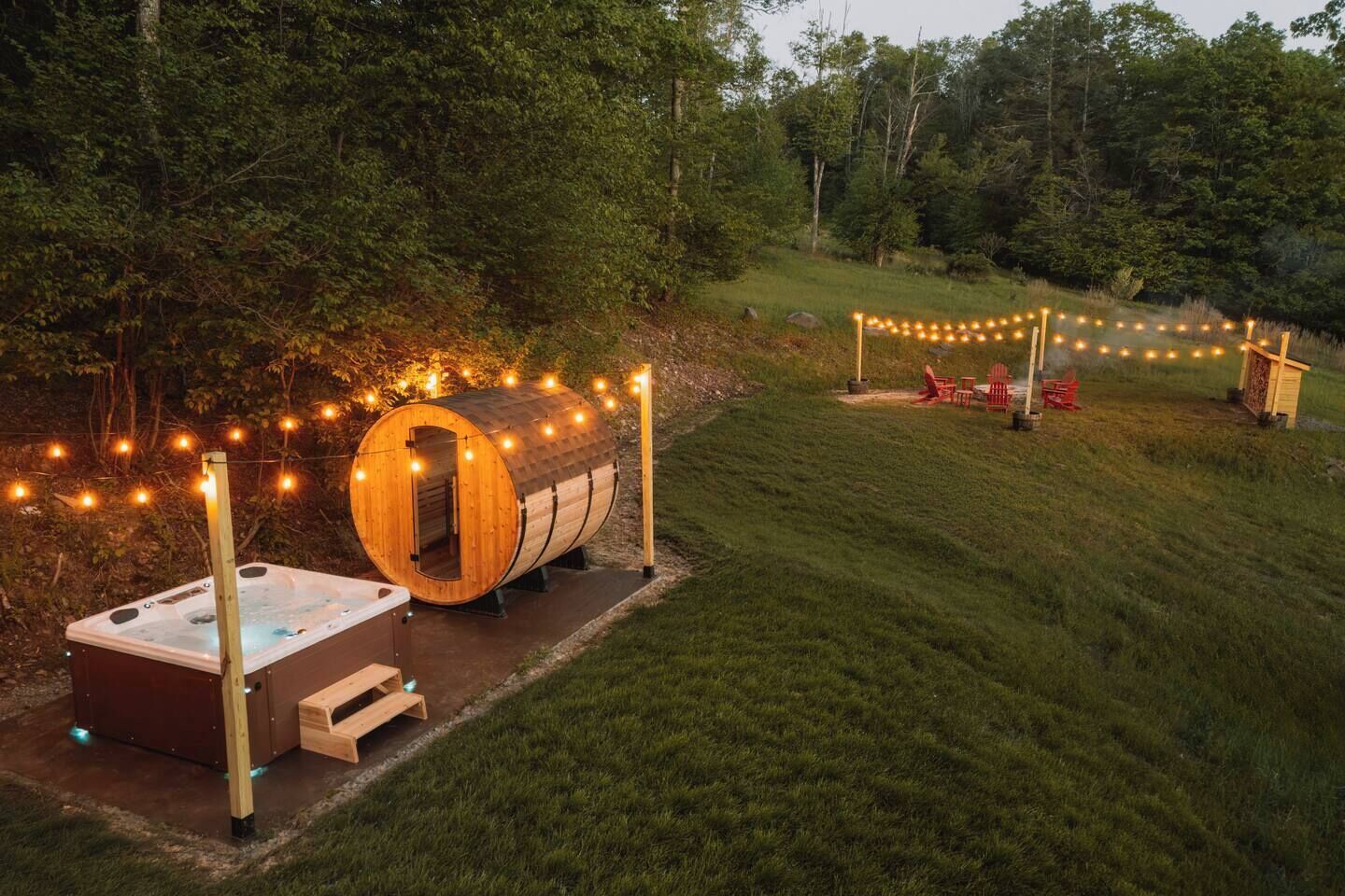 Un bain à remous et un tonneau en bois décorés de guirlandes lumineuses, le tout installé dans un espace extérieur verdoyant.