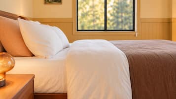 A neatly made bed with white and brown bedding, a bedside table with a lamp, and a window with a view of trees.