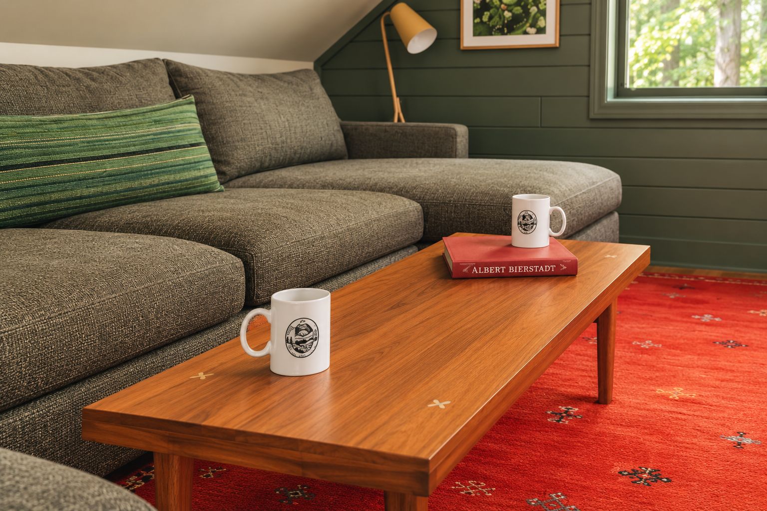 A cozy living room with a wooden coffee table, two white mugs, a red book, and a green patterned cushion on a grey sofa.