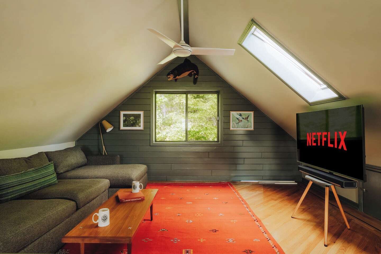 A cozy attic room with a sloped ceiling, a ceiling fan, a large window, a television, a sofa, a coffee table, and a red rug.