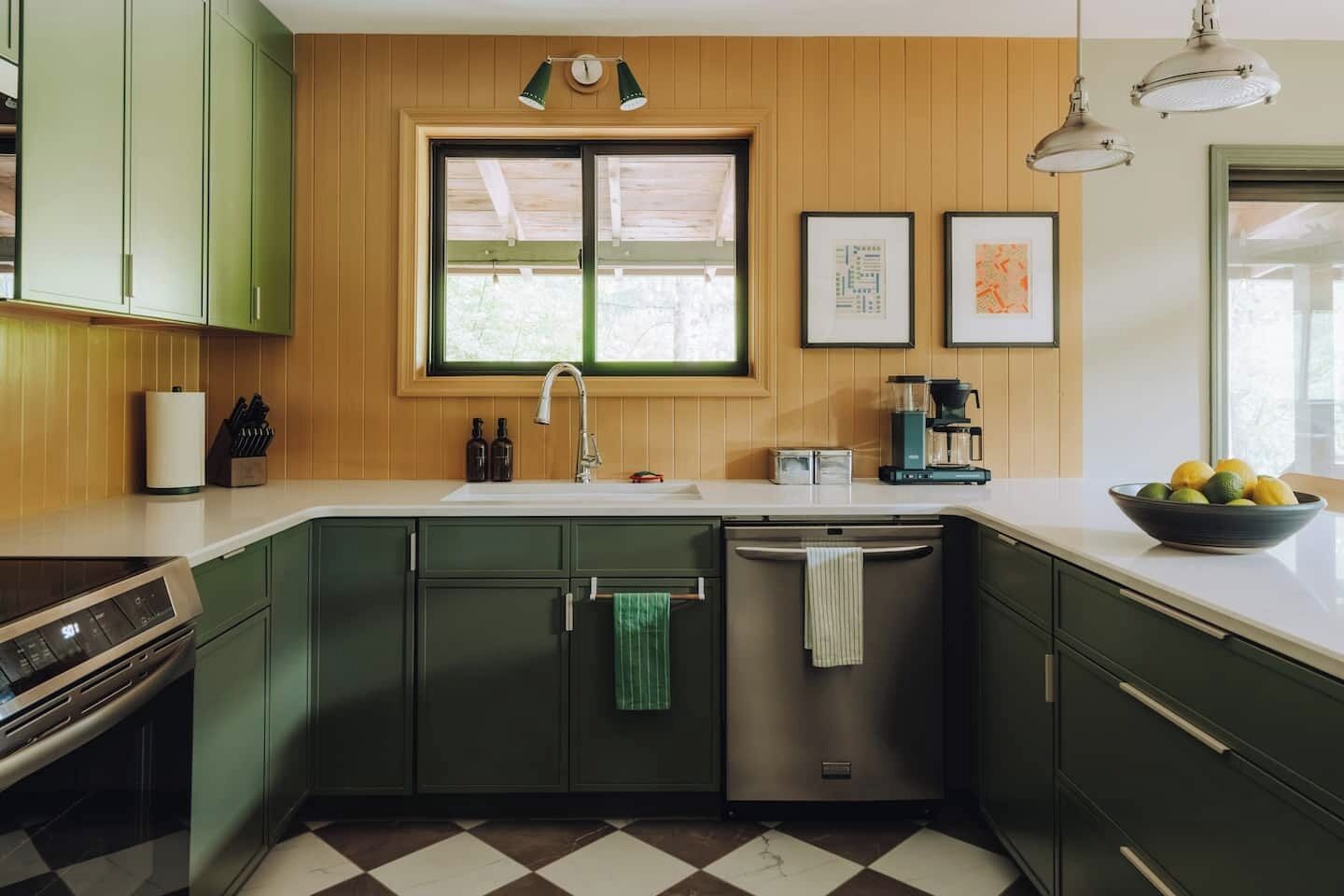 Uma cozinha com armários verdes, uma lava-louças de aço inoxidável, uma cafeteira e uma tigela de frutas na bancada.