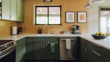 Uma cozinha com armários verdes, uma lava-louças de aço inoxidável, uma cafeteira e uma tigela de frutas na bancada.