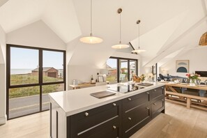 A modern kitchen with a large island, pendant lighting, and a view of the sea. - Seaside House - North East Escapes (Beadnell)