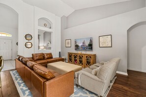 Ein Wohnzimmer mit einem braunen Ledersofa, einem beigen Sessel, einer Holzkommode und einem an der Wand befestigten Fernseher.