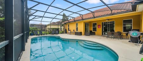 Une piscine intérieure dotée d’un toit en verre, entourée de chaises longues et bordée d’un bâtiment jaune au toit recouvert de tuiles.