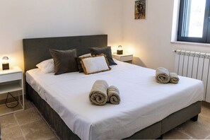 Cama bem arrumada com almofadas e toalhas dobradas, luminária de cabeceira e janela com luz do dia.