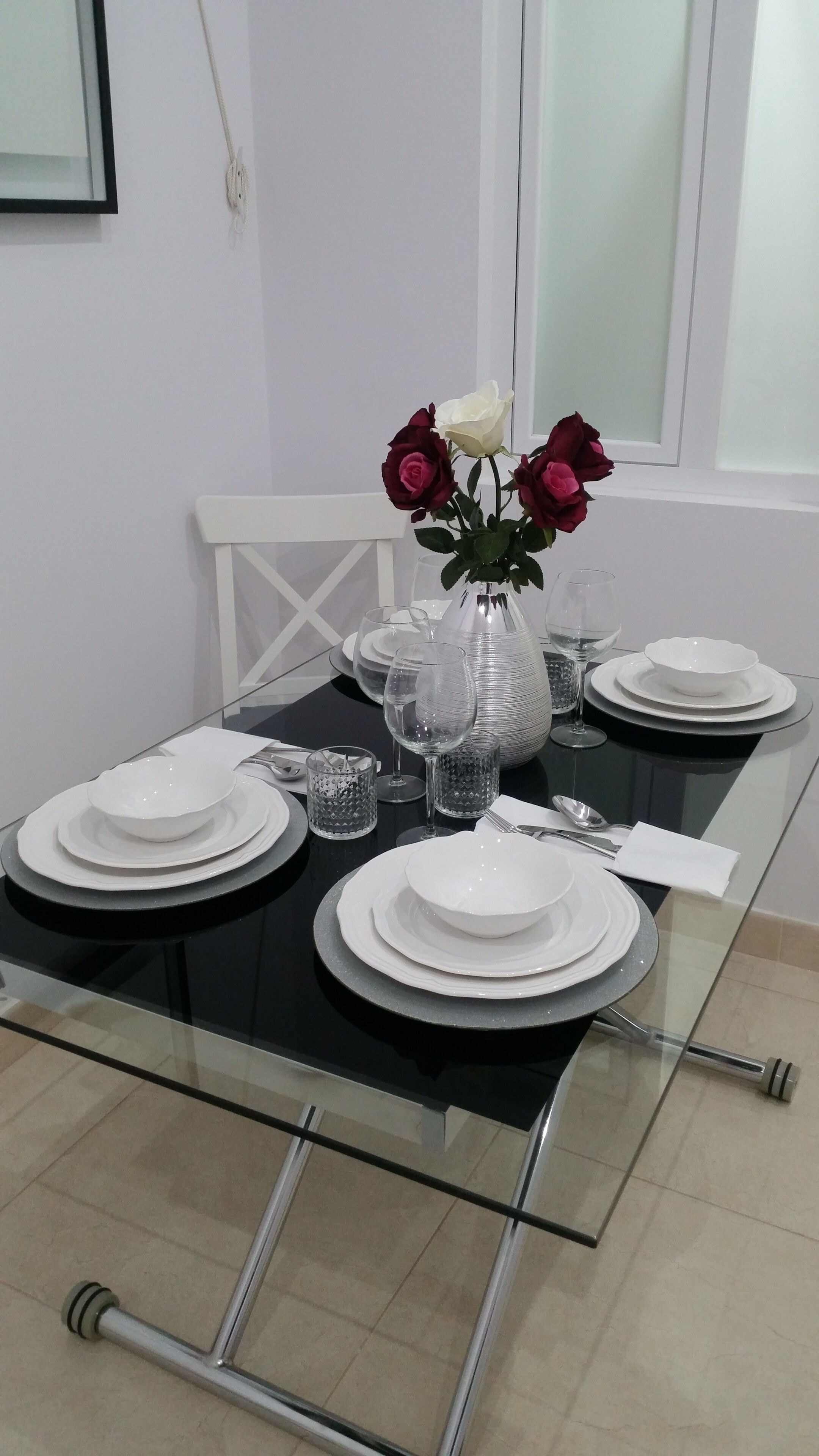 Una mesa de comedor servida con platos blancos, vasos transparentes y un florero con flores.