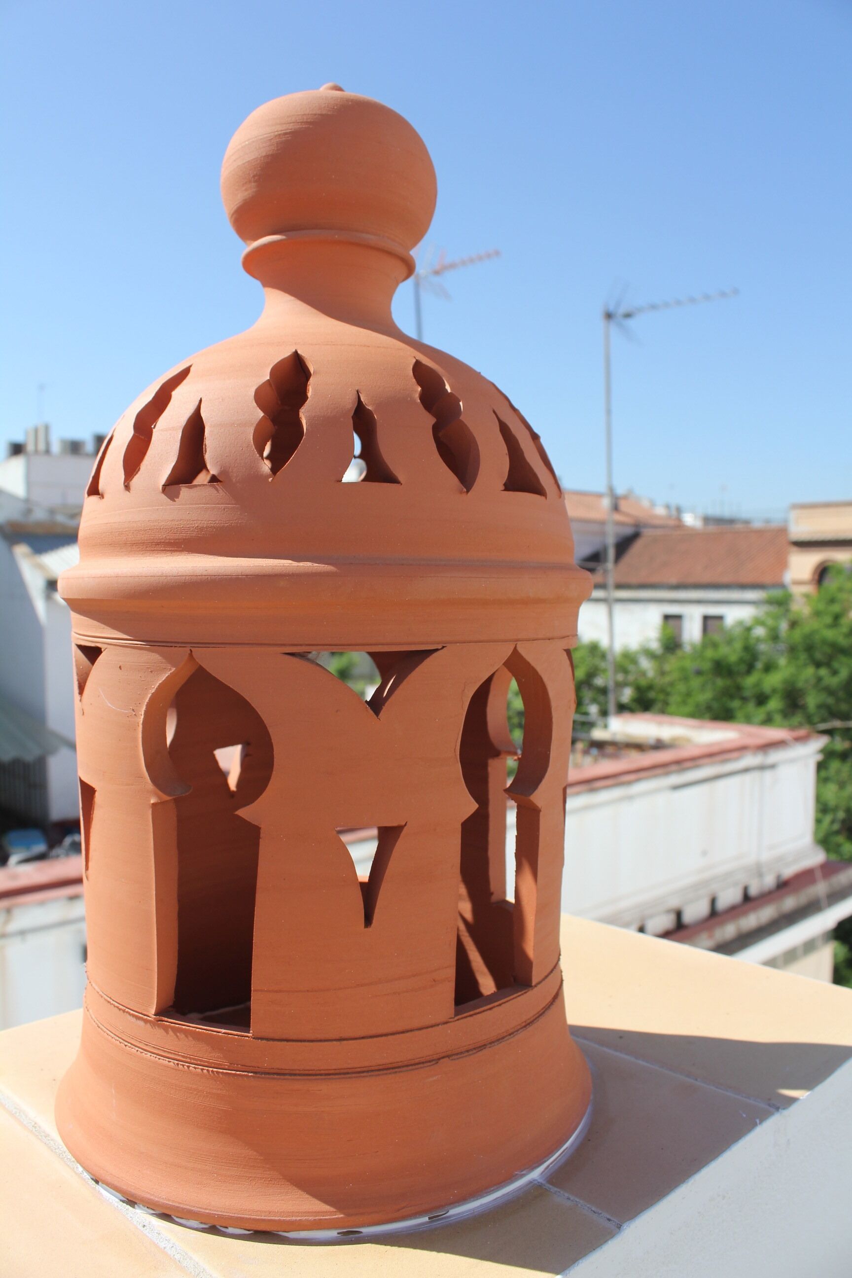 Um candeeiro de terracota com topo arredondado e detalhes recortados numa varanda.