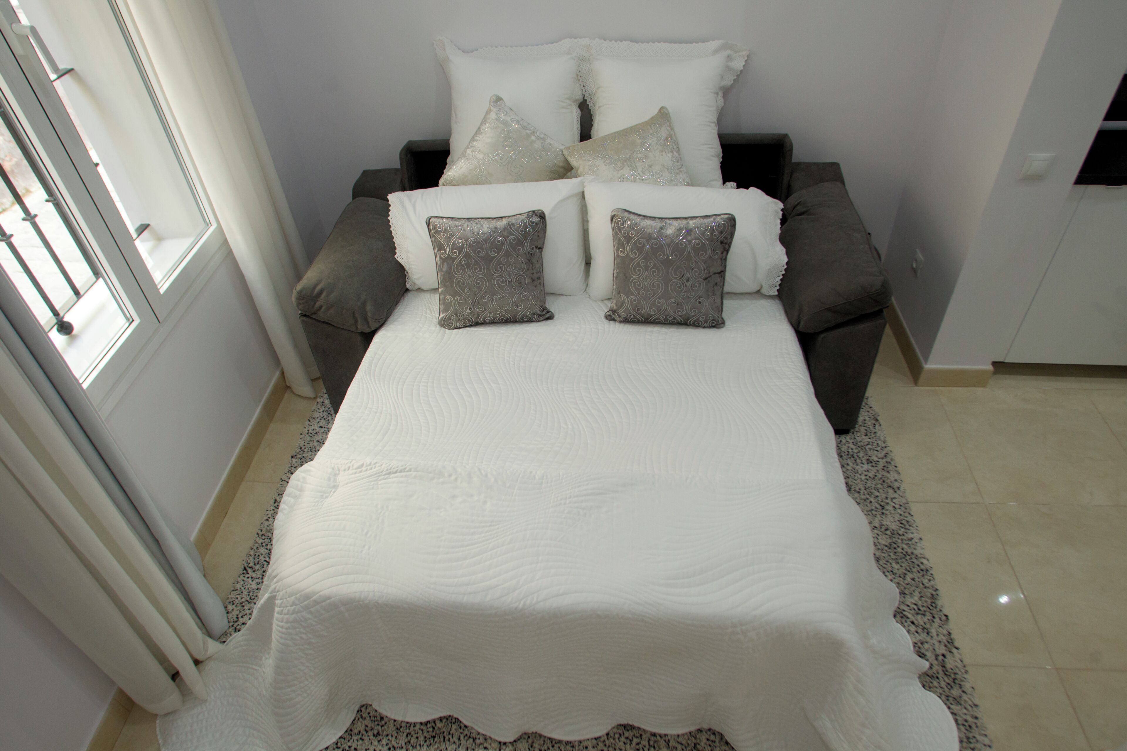 Una cama con una colcha blanca y dos almohadas grises, un sofá gris con una almohada blanca y una silla blanca.