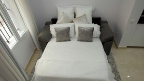 Una cama con una colcha blanca y dos almohadas grises, un sofá gris con una almohada blanca y una silla blanca.