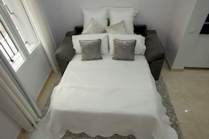 Una cama con una colcha blanca y dos almohadas grises, un sofá gris con una almohada blanca y una silla blanca.