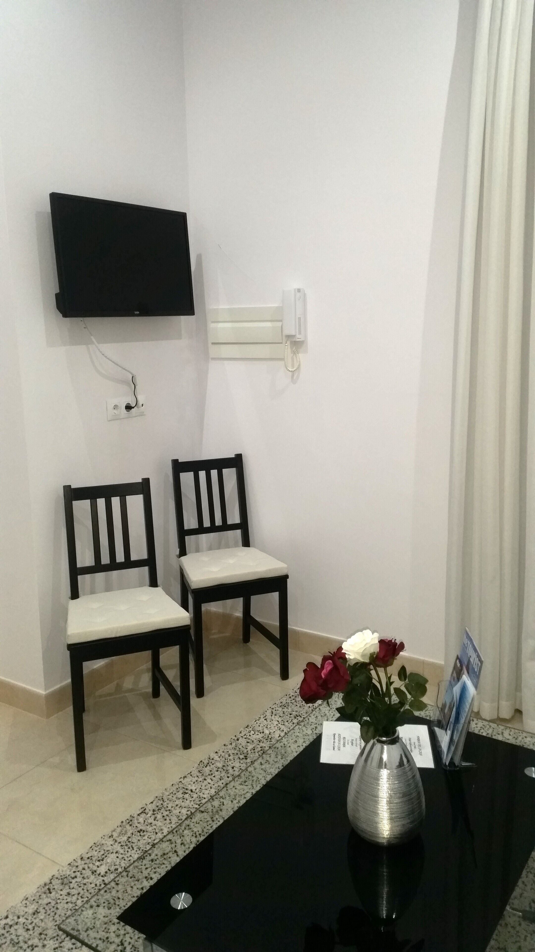 A room with two chairs, a flat-screen TV mounted on the wall, a telephone, a vase with flowers, and a table with a brochure.