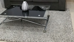 Una sala de estar con un sofá gris, almohadones decorativos, una mesa de centro de vidrio con un florero y un cuadro en la pared.