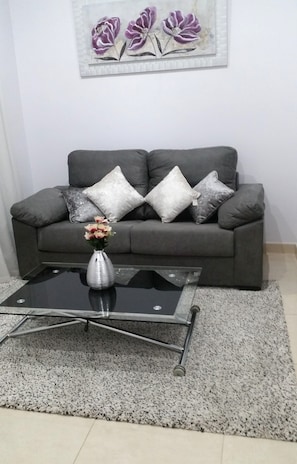 Una sala de estar con un sofá gris, almohadones decorativos, una mesa de centro de vidrio con un florero y un cuadro en la pared.