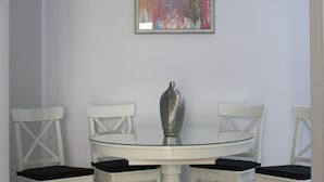 Un comedor con una mesa blanca, cuatro sillas blancas, un florero plateado y un cuadro abstracto colorido en la pared.