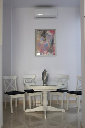 Un comedor con una mesa blanca, cuatro sillas blancas, un florero plateado y un cuadro abstracto colorido en la pared.