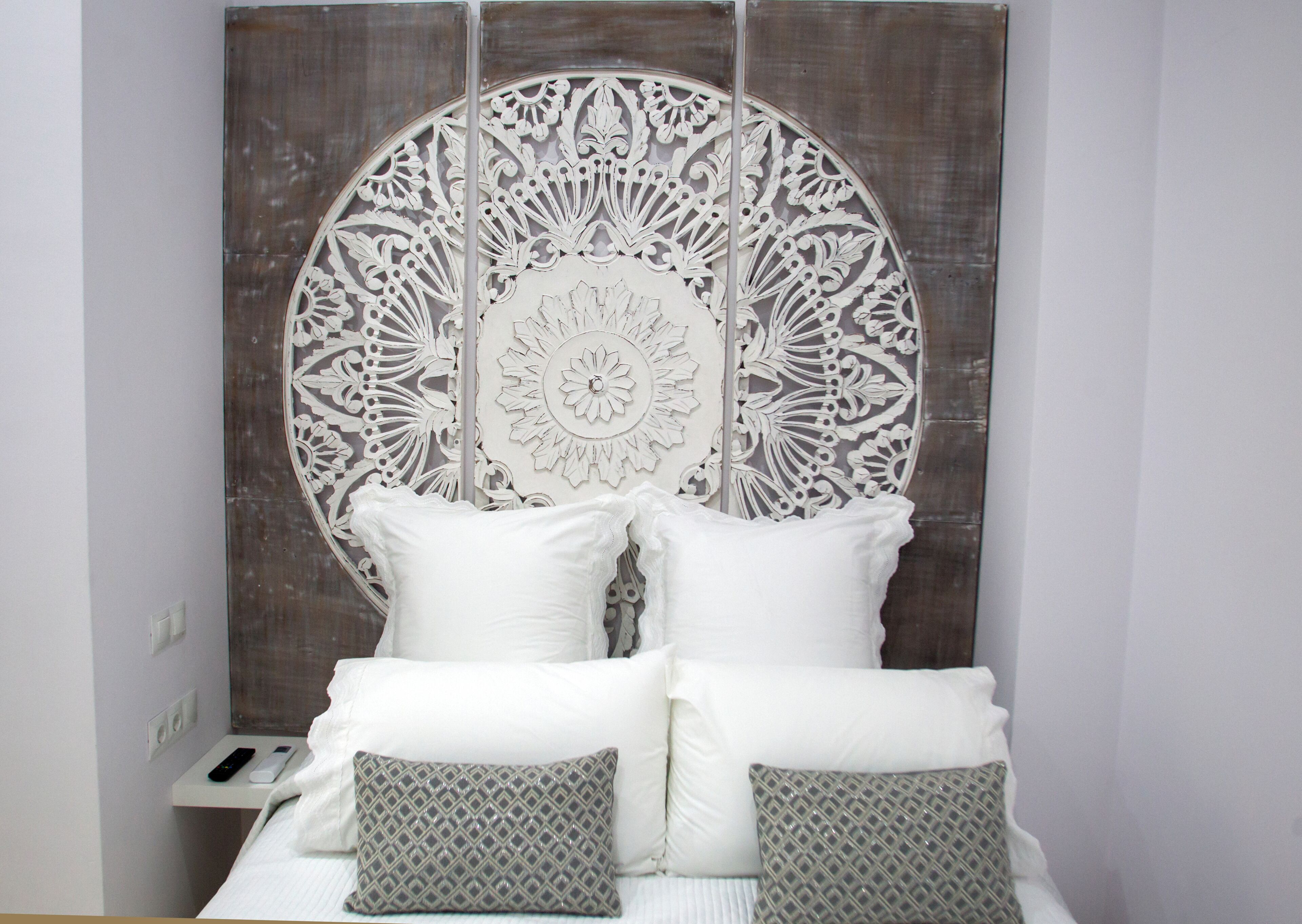 Una cama con ropa de cama blanca y almohadones decorativos, una mesita de noche con un control remoto y una pared con un panel decorativo circular.