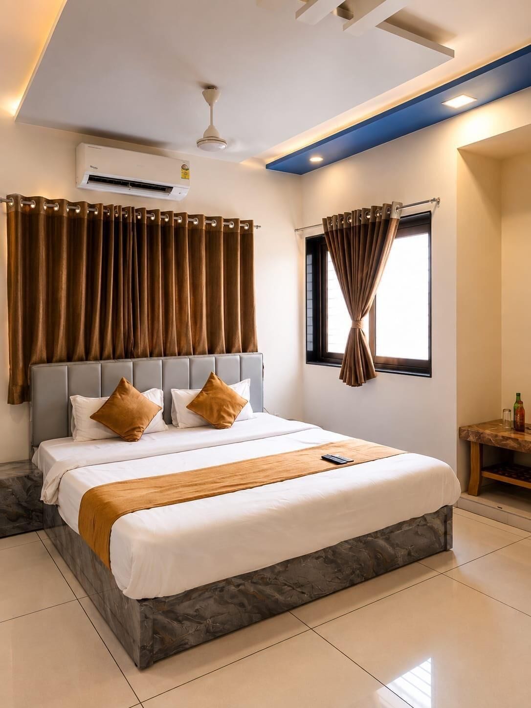 A hotel room with a bed, curtains, a ceiling fan, an air conditioning unit, a window with curtains, and a small table with a bottle.