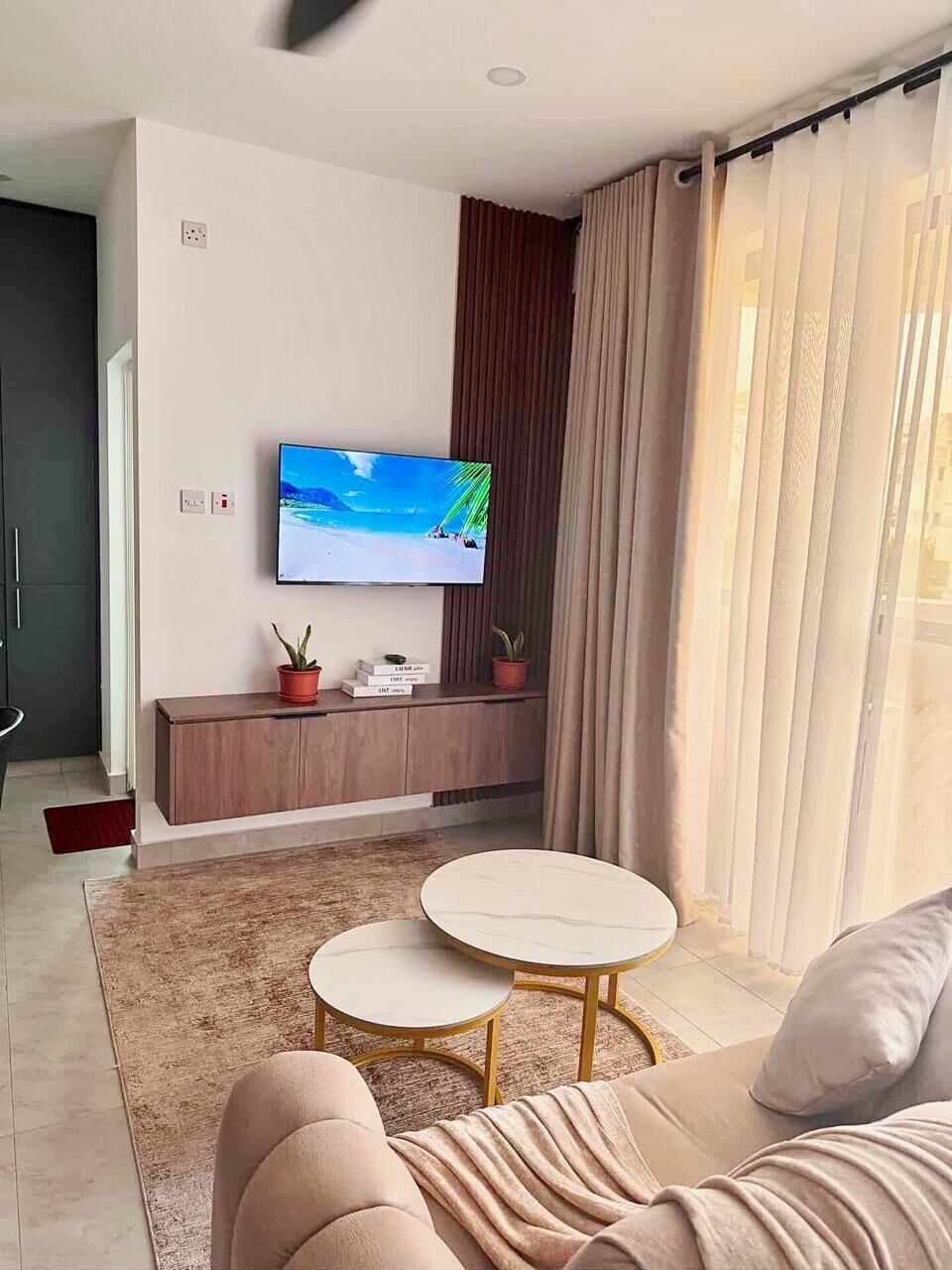 Una sala de estar moderna con un televisor de pantalla plana montado en la pared, un mueble bajo de madera, dos mesas de centro redondas, un sofá color beige y un ventanal con cortinas.