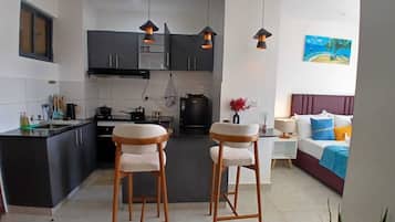 Un espacio habitable compacto con kitchenette, dos taburetes de bar, un sofá y un cuadro de una playa.