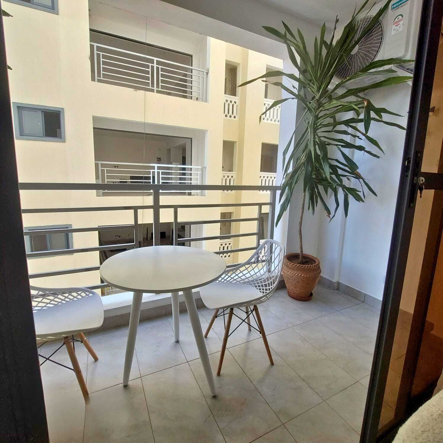 Un balcón con una mesa redonda y sillas, una planta en maceta y vistas de un edificio con balcones.