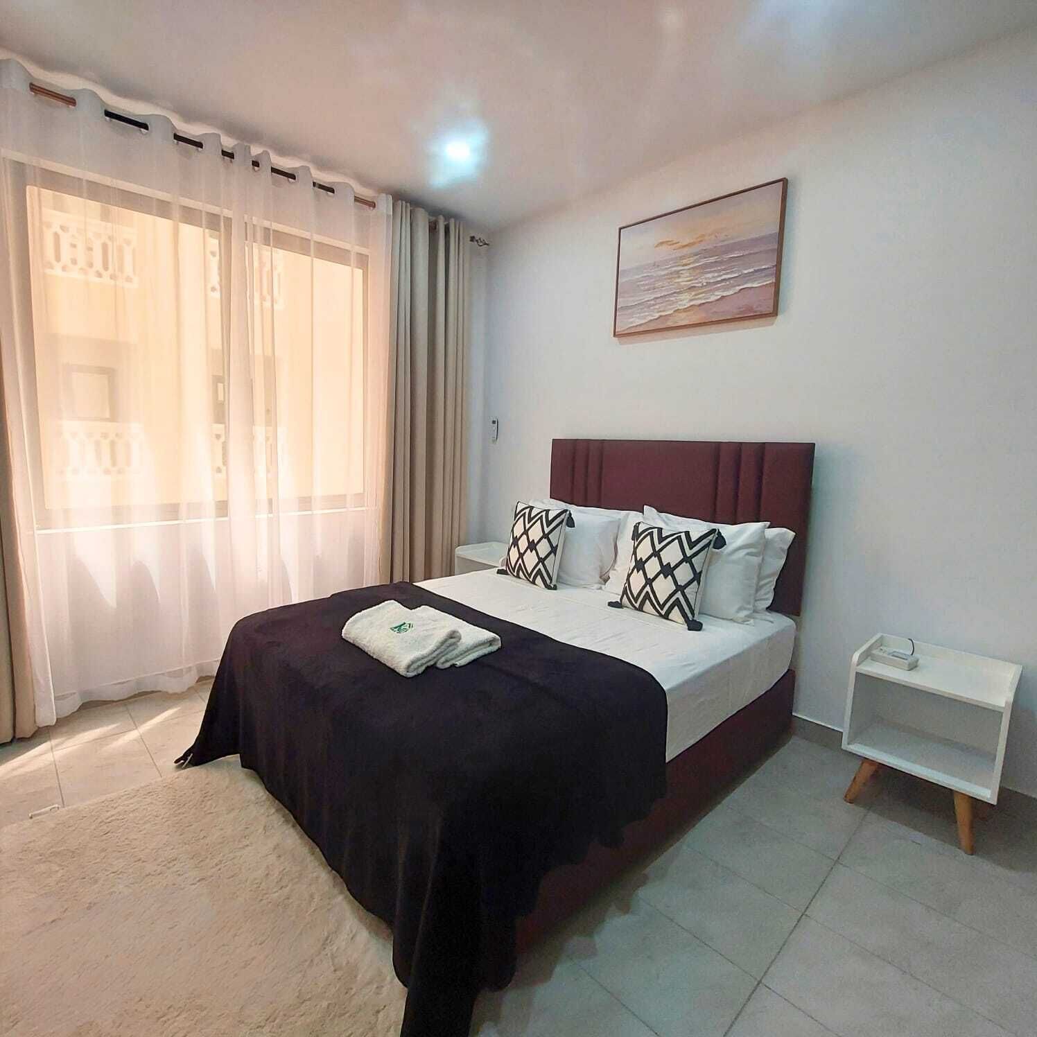 Una habitación de hotel con cama, mesita de noche, un cuadro en la pared y una ventana con cortinas.