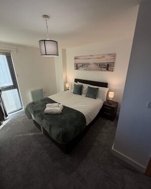 A bedroom with a bed, a nightstand, a lamp, a painting, and a window with blinds. - Elegant 2-Bed w/ Balcony | City Centre (Liverpool)