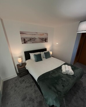 A bedroom with a bed, a nightstand, a lamp, a painting on the wall, and a door. - Elegant 2-Bed w/ Balcony | City Centre (Liverpool)